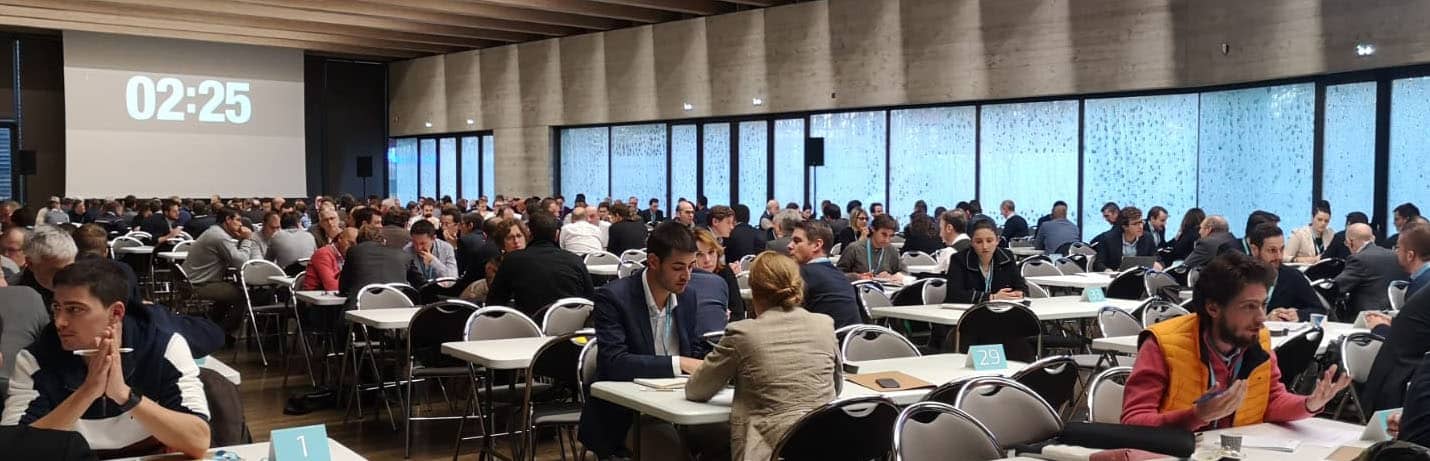Grande salle, nombreux participants autour de tables, compteur visible.